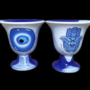 Könnte beinhalten: Zwei weiße Keramikbecher mit blauen und weißen Designs. Ein Becher zeigt ein blaues böses Auge und der andere eine blaue Hamsa-Hand mit dem Wort "Griechenland" darauf.