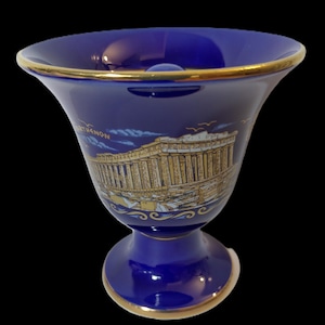 May include: A cobalt blue ceramic vase with a gold rim and base. The vase features a depiction of the Parthenon in Athens, Greece, with gold accents. The vase is a souvenir from Greece.