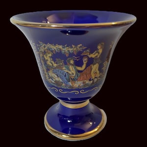 May include: A blue and gold ceramic vase with a decorative design of people and vines. The vase has a wide mouth and a tall, slender base.