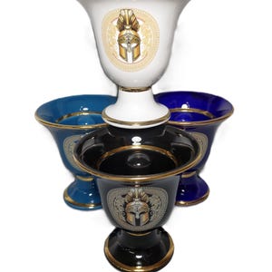 May include: A collection of ceramic goblets in white, blue, and black, each adorned with a gold-trimmed rim and a detailed illustration of a helmet within a circular emblem. The goblets have a pedestal base.