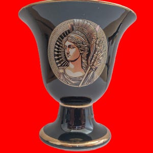 Könnte beinhalten: Schwarze Keramikvase mit goldenem Rand und Sockel. Die Vase zeigt ein rundes Medaillon mit einem klassischen weiblichen Profil, das einen Stab hält. Das Medaillon ist von einem dekorativen Rahmen umgeben.