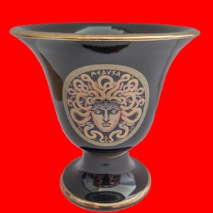 Peut inclure: Vase en céramique noire avec un bord doré et une image dorée et blanche de Méduse avec des serpents pour cheveux. Le vase porte l'inscription "MEDUSA" en or sur le devant.