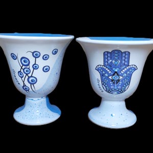 Peut inclure: Deux tasses en céramique blanche avec des motifs bleus et blancs. Une tasse présente un motif bleu de mauvais œil avec le mot "Grèce" écrit sur le côté. L'autre tasse présente un motif bleu de main de Hamsa avec le mot "Grèce" écrit sur le côté.