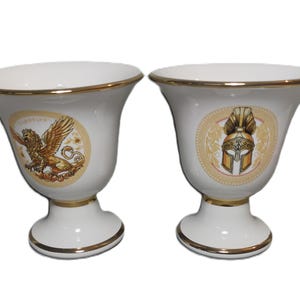 May include: Two white ceramic vases with gold trim. One vase features a griffin design, the other a golden helmet. The vases have a flared top and a pedestal base, suitable for decorative use.