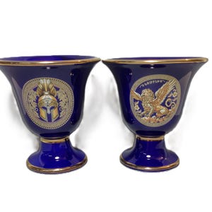 May include: Two blue and gold ceramic cups with decorative designs. One cup features a golden helmet and the other features a golden griffin.