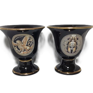 May include: Two black ceramic vases with gold trim. One vase features a gold winged lion with the text "Griffin" and the other vase features a gold helmet with a gold laurel wreath.