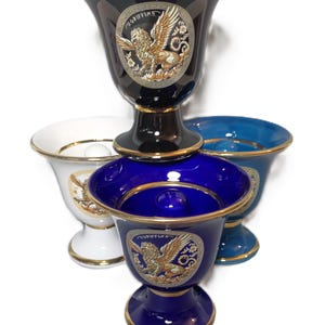 Puede incluir: Colección de jarrones decorativos de cerámica en varios colores: azul marino, blanco y verde azulado. Cada jarrón presenta un borde dorado y un emblema detallado de un grifo. Los jarrones están apilados, mostrando sus diferentes formas y tamaños.