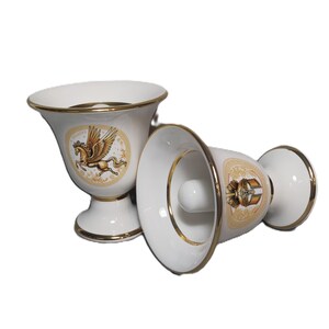 May include: Two white ceramic cups with gold trim. One cup features a winged horse design and the other has a gold emblem. Both cups have a pedestal base.