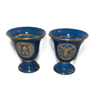 May include: Two blue ceramic cups with gold trim and decorative designs. One cup features a gold helmet design and the other features a gold angel design.