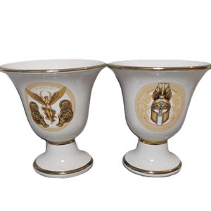 May include: Two white ceramic cups with gold trim and decorative designs. One cup features a winged figure with a golden halo, while the other cup features a golden helmet with a plume.