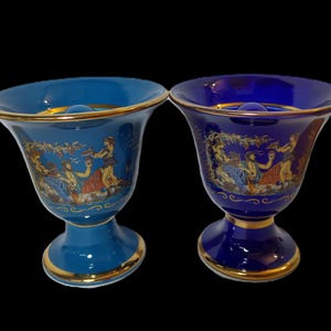 May include: Two blue ceramic vases with gold trim and pedestal bases. Each vase features a classical scene with figures in various colors. The vases have a flared rim and a small, round indentation near the top.
