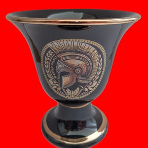 Könnte beinhalten: Schwarzer Keramikkelch mit goldfarbenem Rand und einem goldenen und kupferfarbenen Design eines spartanischen Helms mit Lorbeerkranz. Das Design enthält den Text "SPARTAN HELMET".