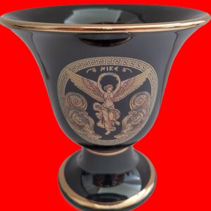 May include: Black ceramic vase with a gold rim and a gold Greek-style design featuring a winged figure of Nike, the Greek goddess of victory. The design includes the word "NIKE" in gold lettering.