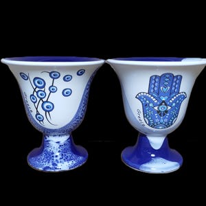 Könnte beinhalten: Zwei weiße Keramikbecher mit blauen und weißen Designs. Der Becher links hat ein Design mit blauen bösen Augen auf einem weißen Stiel. Der Becher rechts hat eine blaue Hamsa-Hand mit einem bösen Auge in der Mitte. Beide Becher haben das Wort "Griechenland" darauf geschrieben.