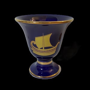 May include: A cobalt blue ceramic vase with a gold trim and a gold image of a ship with a sail. The vase has a pedestal base.