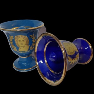 Pode incluir: Duas xícaras de cerâmica azul e dourada com uma borda dourada. Uma xícara apresenta uma imagem dourada de Alexandre, o Grande, com uma coroa de louro. A outra xícara tem uma imagem dourada da cabeça de uma pessoa com uma coroa de louro.
