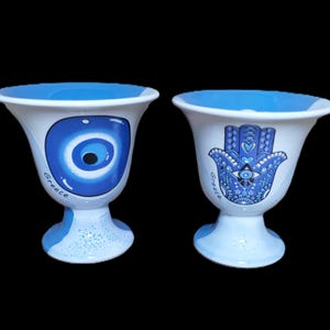 May include: Two white ceramic goblets with blue rims and bases. One goblet features a blue evil eye design, the other a blue Hamsa hand. Both have the word "Greece" in script.