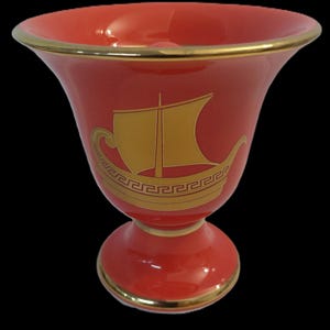 May include: A red ceramic vase with a gold trim and a gold ship design. The ship has a Greek key border.