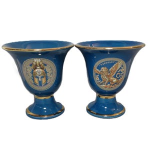 May include: Two blue ceramic vases with gold trim and decorative designs. One vase features a gold helmet and the other features a gold griffin.