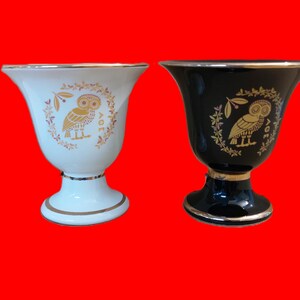Puede incluir: Dos tazas de cerámica con ribetes dorados y un diseño de búho. La taza de la izquierda es blanca con un búho dorado y el texto "AOE". La taza de la derecha es negra con un búho dorado y el texto "LOV".
