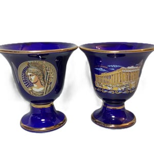 May include: Two blue and gold ceramic cups with decorative designs. One cup features a profile of a woman with a crown and a spear. The other cup features a gold building with the text "ATHENON" above it.