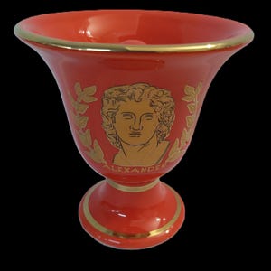 Puede incluir: Un gobelet de cerámica roja con un borde y una base dorados. El gobelet presenta una imagen dorada de Alejandro Magno con hojas de laurel.