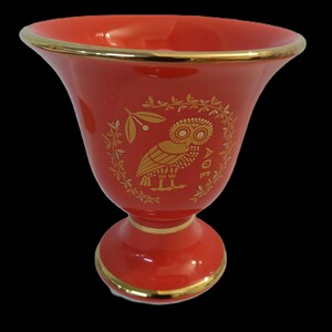 May include: A red ceramic vase with a gold rim and a gold owl design. The owl is surrounded by a wreath of leaves and the word "AOE" is written below it.