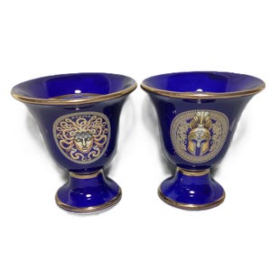 May include: Two blue ceramic vases with gold trim. One vase features a gold image of Medusa, the other features a gold image of a Spartan helmet.