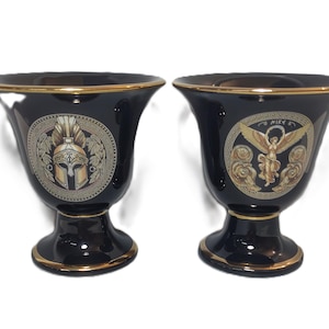 May include: Two black ceramic vases with gold trim and decorative designs. One vase features a gold helmet with a plume, the other features a gold winged figure with the word "NIKE" above it.