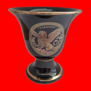 Könnte beinhalten: Schwarze Keramikvase mit goldener Verzierung und einem goldenen Bild eines Greifens mit dem Wort "GRIFFING" in einem Kreis darum.