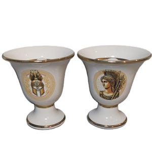 May include: Two white ceramic cups with gold trim and a design of a Greek warrior and a Greek goddess. The cups are on a white background.