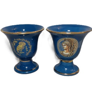 May include: Two blue ceramic vases with gold trim. One vase features a gold owl design with the text "AOE", and the other has a gold illustration of a woman. The vases have a flared top and a pedestal base.