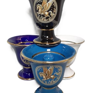 May include: A collection of ceramic shaving mugs in various colors: navy blue, white, and light blue. Each mug features a gold rim and a Pegasus emblem. The mugs are stacked, showcasing their design and shape.