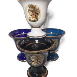 Puede incluir: Tres tazas de cerámica con ribetes dorados y un retrato de una mujer con casco. Las tazas son de color negro, blanco y azul.