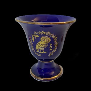 以下が含まれることがあります： 金色の縁取りと金色のフクロウのデザインが施された青いセラミック製のゴブレット。フクロウはオリーブの枝の輪で囲まれており、その下に「AOE」という言葉が書かれている。