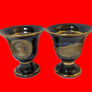 May include: Two black ceramic cups with gold trim and painted designs. One cup features a woman's face and the other features a building.