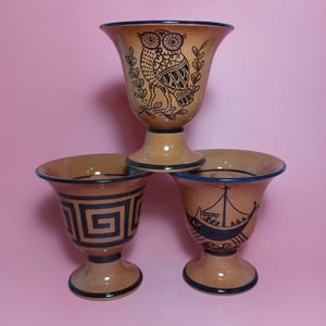 May include: Three brown ceramic cups with black and blue painted designs. The top cup features an owl, the middle cup has a geometric pattern, and the bottom cup has a ship with people on it.