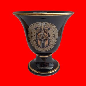 May include: Black ceramic goblet with a gold rim and a gold design of a Spartan helmet with a plume on a black background.