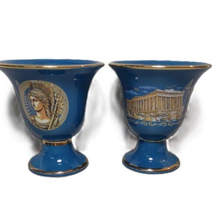 May include: Two blue ceramic cups with gold trim. One cup features a depiction of a woman with a helmet and a spear, while the other cup features a depiction of a building with columns.