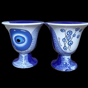 May include: Two blue and white ceramic cups with a Greek evil eye design. One cup features a single large evil eye, while the other has a vine of smaller evil eyes. The word "Greece" is written on the cup with the vine of evil eyes.
