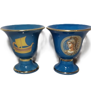 May include: Two blue ceramic vases with gold trim. One vase features a yellow ship with red and gold details. The other vase features a woman with a gold crown and a spear.