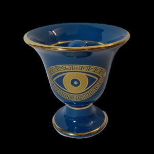 Puede incluir: Un jarrón de cerámica azul con un borde dorado y un diseño de ojo dorado con un patrón de llave griega. El jarrón tiene una base redonda.