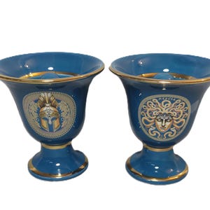 May include: Two blue ceramic cups with gold trim and decorative designs. One cup features a gold helmet design and the other features a gold Medusa design.