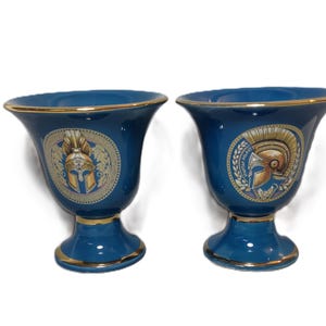 May include: Two blue ceramic cups with gold trim and a design of a gold helmet with a red plume on a white background. The cups are on a white background.