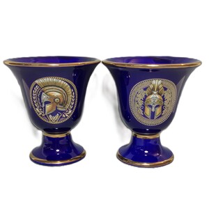 Könnte beinhalten: Zwei blaue und goldene Keramikvasen mit einem griechischen Kriegerhelm-Design. Die Vasen haben eine breite Öffnung und einen schmalen Boden.