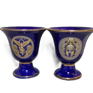 May include: Two cobalt blue ceramic vases with gold trim. Each vase features a detailed gold emblem. One depicts a winged figure with the word "NIKE", the other a helmet. The vases have a pedestal base.