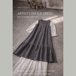 Könnte beinhalten: Ein graues Artist's Smock Kleid mit einem weißen Unterhemd, mit V-Ausschnitt und Knopfleiste. Das Kleid hat einen gerüschten Saum und ist ein PDF-Schnittmuster. Der Text auf dem Bild lautet: "ANNA VEJS STUDIO" und "ARTIST'S SMOCK DRESS PDF SEWING PATTERN".
