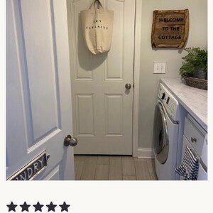 Pu&ograve; includere: Una borsa per la biancheria in tela beige appesa a una porta. La borsa ha un testo nero che recita "DRY CLEANING". Un cartello di legno con la scritta "WELCOME TO THE COTTAGE" &egrave; montato sulla parete.