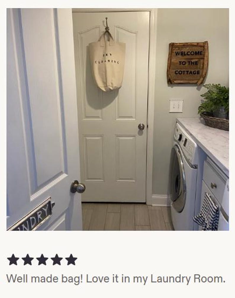 Pu&ograve; includere: Una borsa per la biancheria in tela beige appesa a una porta. La borsa ha un testo nero che recita "DRY CLEANING". Un cartello di legno con la scritta "WELCOME TO THE COTTAGE" &egrave; montato sulla parete.