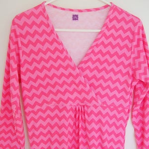 May include: Pink long-sleeved shirt with a chevron pattern and a V-neck. The shirt has a gathered waist and a small tag that says "Lily".